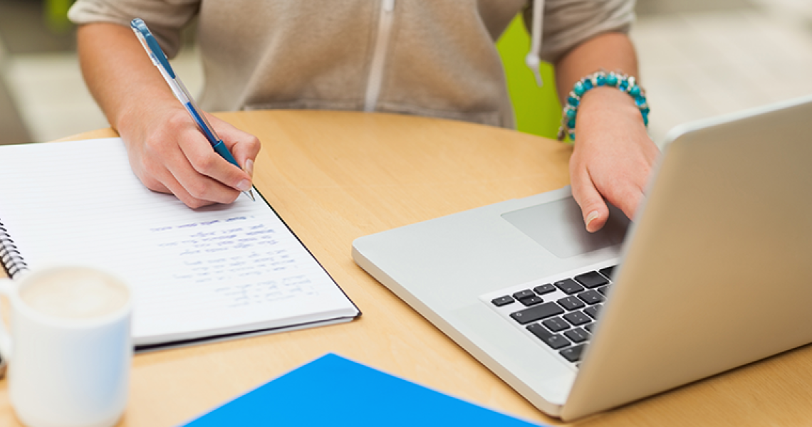 Close up of hand writing in a notebook, next to a laptop